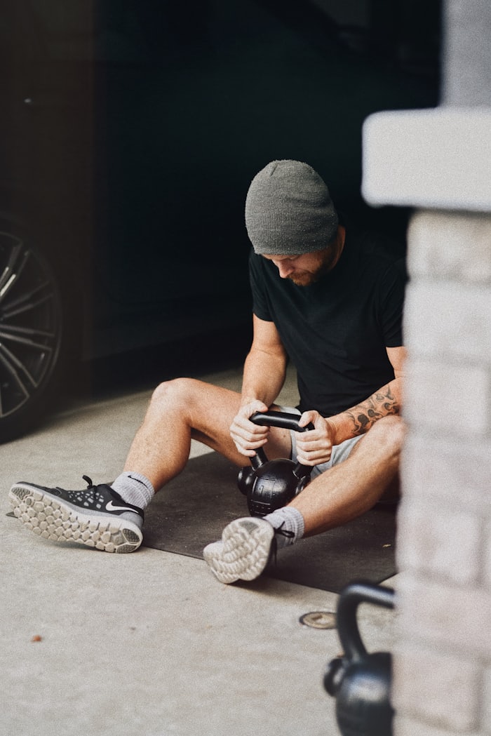 A man in a beanie sitting in his garage with a kettlebell