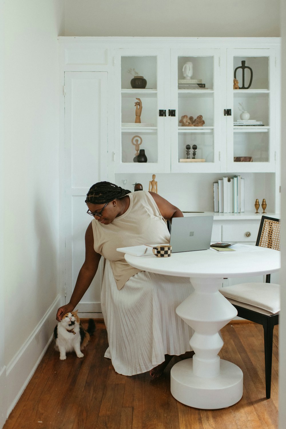 A woman at her table at home reaching down to pet her cat