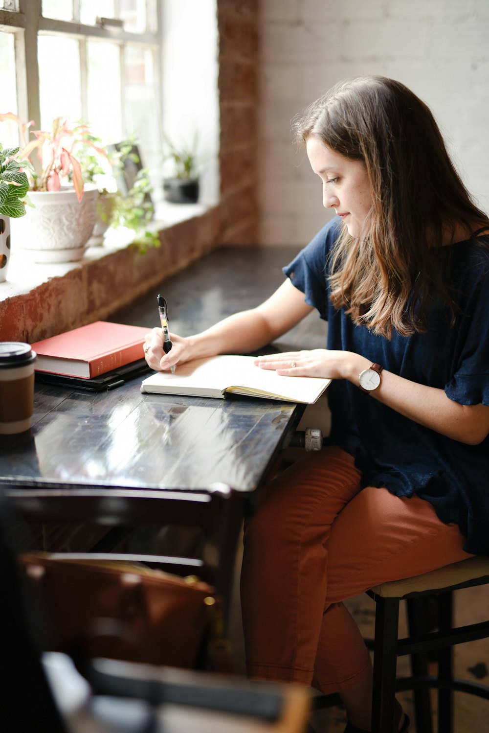 A woman writes in a journal at a table beside a bright window lined with plants